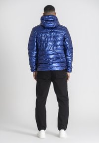 Blå vadderad jacka med en blank yta, försedd med huva och horisontella sömdekorationer. Bärs över svarta byxor och vita sneakers.