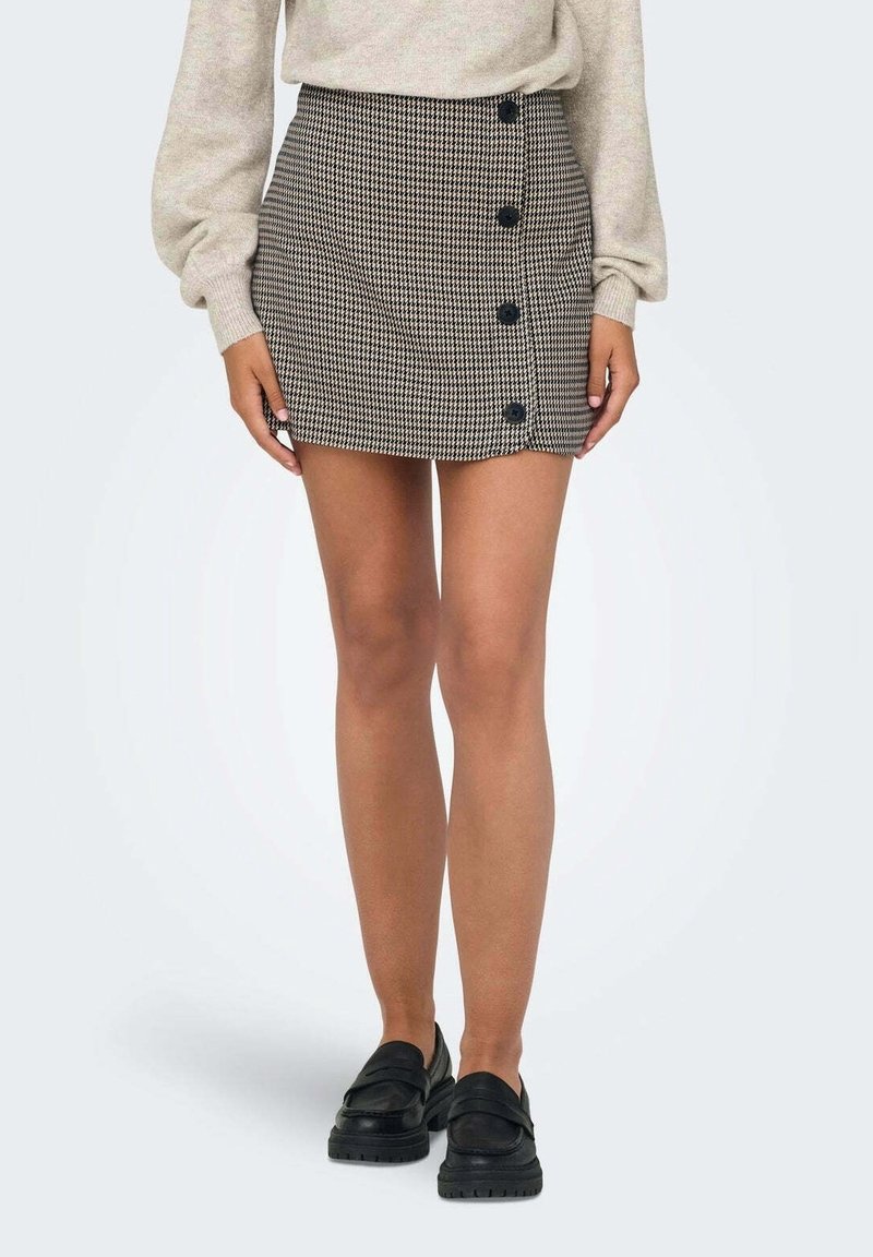 Femme portant un pull beige, une mini-jupe pied-de-poule avec des boutons noirs sur le côté, et des mocassins noirs, debout les bras détendus.