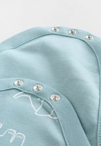 Hele sinine kangas sujuva tekstuuriga, millel on hõbedased nööbid ja peen valge graafiline disain. Ümar kaeluseäär on nähtav.