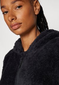 Schwarze, flauschige Zip-Hoodie mit weicher Textur und Kapuze. Hat einen Reißverschluss und einen runden Ausschnitt, vor einem weißen Hintergrund.