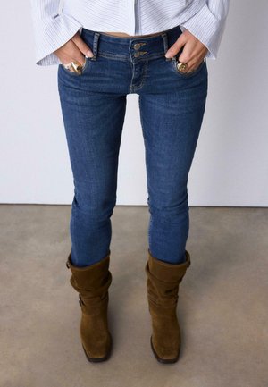 Personne portant un jean skinny bleu, des bottes marron en daim souples, une chemise blanche à rayures et des bijoux aux mains avec les doigts dans les poches.