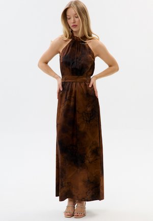 Donna che indossa un vestito lungo senza maniche con stampa tie-dye marrone e nera, collo all'americana e cintura, in piedi con le mani sui fianchi su uno sfondo bianco.