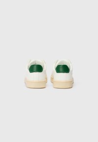 Baskets blanches avec des accents verts au niveau du talon, semelle en caoutchouc beige plate et doublure en tissu texturé. Design minimaliste, forme arrondie.