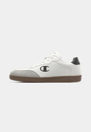 Baskets blanches avec une tige en cuir lisse, des accents en daim gris, un logo noir et une semelle en caoutchouc brun, présentant des finitions cousues et des lacets ronds.