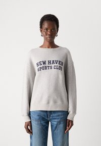 Pilkas džemperis su tamsiai mėlynu tekstu "NEW HAVEN SPORTS CLUB", apvaliu iškirpimu, nusileidusiomis rankovėmis ir nubuksuotais rankogaliais. Derinamas su mėlynomis džinsomis.