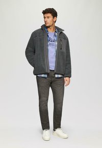 Harmaa fleece-takki korkealla kauluksella, jossa on etuvetoketjut ja sivutaskut, käytetään sinisen T-paidan, ruudullisen paidan ja tummien farkkujen päällä.