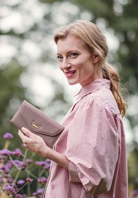 Light pink clutch wallet in textured material, featuring a gold logo accent, held by a person in a soft pink blouse, surrounded by flowers.