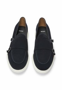 Navy suede slip-on shoes with a wide strap and silver buckle detail. White rubber sole. Interior features a brown lining. Brand logo visible.