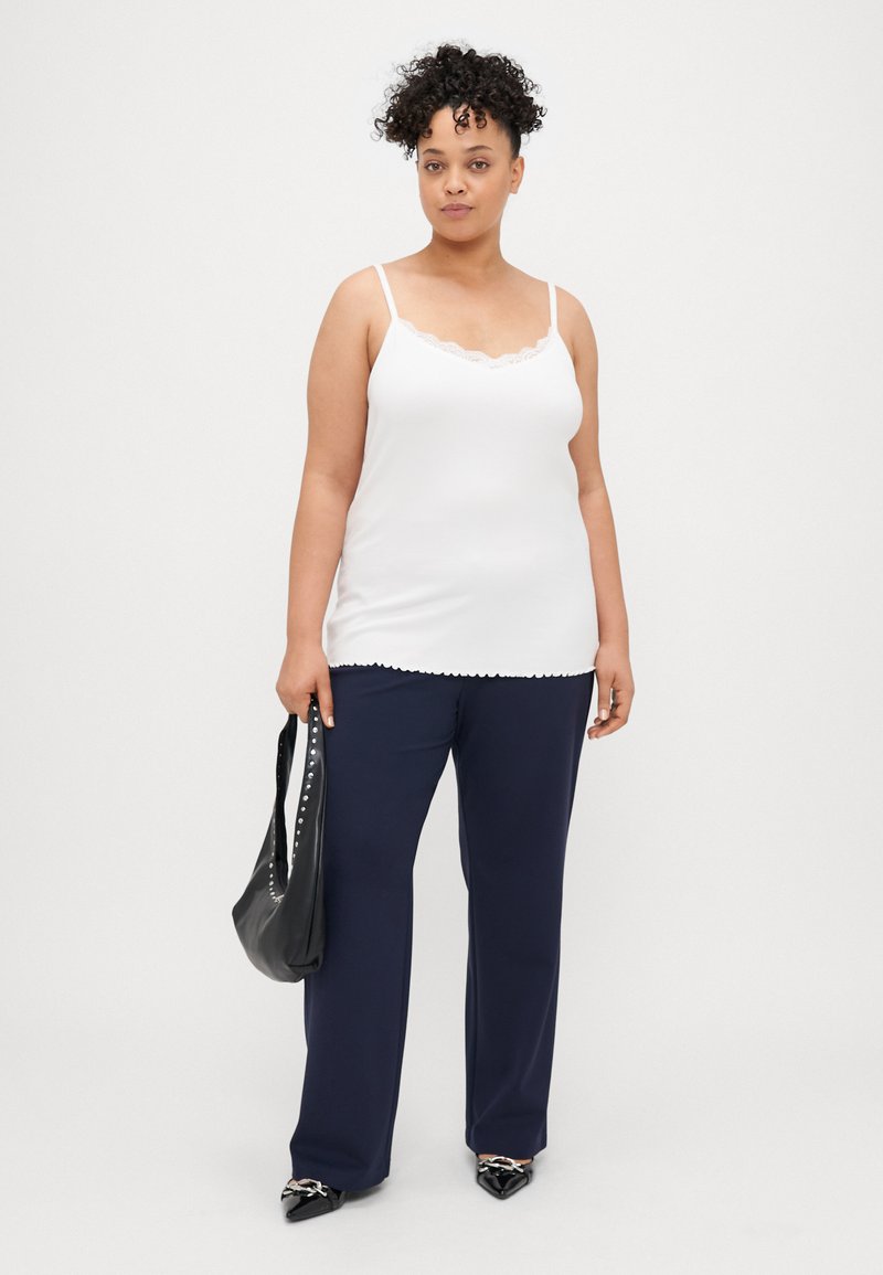 Camisola branca com rendas, combinada com calças largas azul-marinho. A modelo segura uma mala preta com detalhes de tachas.