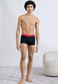 Μαύρα μπόξερ εσώρουχα με κόκκινη μέση που φέρει λογότυπο. Έχουν στενή εφαρμογή και λεία υφή. Το μοντέλο στέκεται μπροστά σε ουδέτερο φόντο.