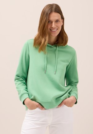 Felpa verde chiaro con tasca a marsupio frontale, lacci e polsini a coste. Abbinata a pantaloni bianchi per un look informale.