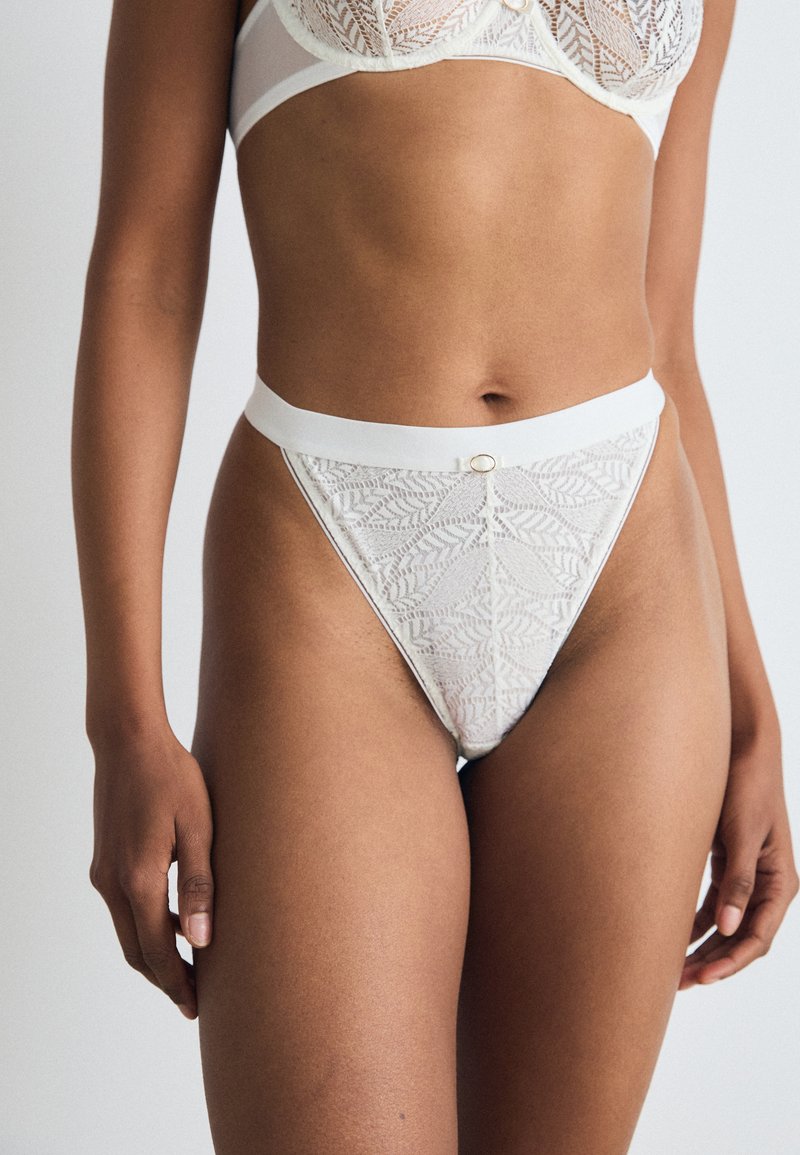 Weiße Spitzen-Tanga mit floralem Muster und glattem Bund. Hat eine kleine goldene Akzentuierung an der Vorderseite. Weicher Stoff, minimales Abdeckungsdesign.