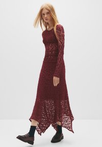 Burgunderfarbenes langärmeliges Spitzenkleid mit strukturiertem Muster, asymmetrischem Saum und durchscheinendem unteren Teil, kombiniert mit dunklen Schuhen und Socken.