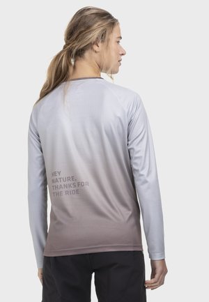 Frau mit blondem Pferdeschwanz trägt ein langärmeliges Shirt mit Farbverlauf von Hellgrau zu Braun und dem Text "HEY NATURE, THANKS FOR THE RIDE" auf dem Rücken.
