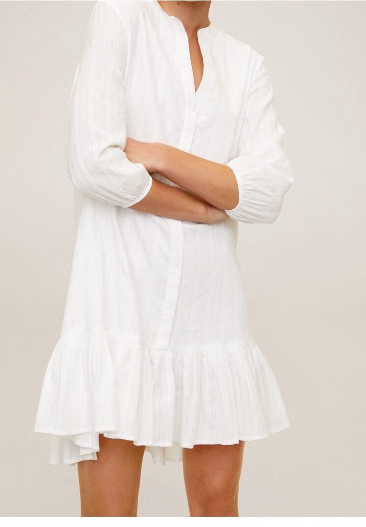 Femme portant une robe blanche à manches longues et longueur genoux avec un ourlet à volants, debout les bras croisés devant un fond uni.