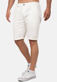 Homme portant un short blanc retroussé et des baskets blanches à faible hauteur avec des chaussettes basses, se tenant devant un fond uni.
