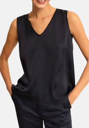 Donna che indossa una camicetta nera senza maniche con scollo a V, mani nelle tasche, sorridente su uno sfondo chiaro e uniforme.