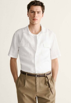 Joven con camisa blanca de mangas cortas abotonada metida en pantalones beige con cinturón trenzado marrón, manos en los bolsillos.