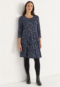 Sininen mekko, jossa on musta pystysuuntainen kuvio, pyöreä pääntie, kolmannes pitkä hiha, polven pituinen, yhdistetty mustiin legginsseihin ja nilkkabootseihin.