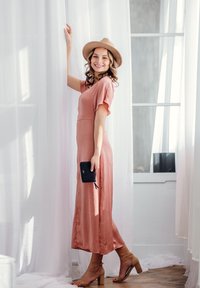 Light pink, knee-length dress with short sleeves, paired with beige block-heel sandals. Black clutch in hand, standing by sheer curtains.