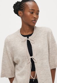 Beige knitted cardigan with short sleeves, featuring a tied front closure and a soft texture, layered over a black top.