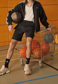 Person i sportkläder som håller en basketboll och lutar sig mot en metallvagn fylld med basketbollar i en inomhushall.