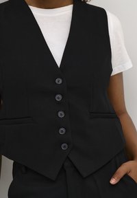 Gilet nero su misura con design aderente, cinque bottoni neri e due tasche frontali, abbinato a una t-shirt bianca sotto.