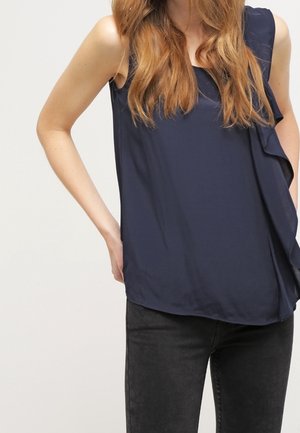 Vrouw die een mouwloze marineblauwe blouse met ruches en een zwarte spijkerbroek draagt, poseert met één hand op haar heup tegen een effen achtergrond.