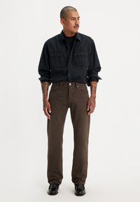 Camisa de mezclilla oscura con dos bolsillos en el pecho, combinada con pantalones rectos marrones. Zapatos negros completan el conjunto. Diseño y ajuste minimalista.