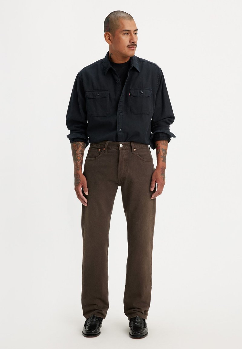 Camisa de mezclilla oscura con dos bolsillos en el pecho, combinada con pantalones rectos marrones. Zapatos negros completan el conjunto. Diseño y ajuste minimalista.