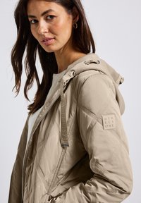 Gequilted beige jas met een capuchon, voorzien van een merkaccent op de mouw, metalen rits en textuurpatronen op de stof.