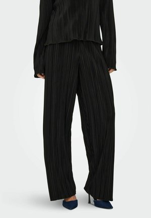 Černý plisovaný outfit se volným topem a širokými kalhotami, vyrobený z jemného materiálu s texturovaným povrchem. Doplněný o špičaté lodičky.