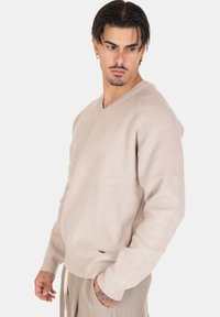 Maglione beige a coste con scollatura a V, maniche lunghe, vestibilità rilassata e un piccolo logo decorativo. Texture morbida e design informale.