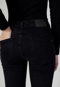 Jeans in denim nero con un design aderente, dotati di tasche posteriori e un'etichetta in vita. La texture del tessuto appare liscia con una leggera scoloritura.