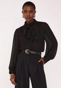 Blouse noire transparente avec un détail de col noué et des manches longues, associée à un pantalon noir taille haute et une ceinture noire texturée avec une boucle dorée.
