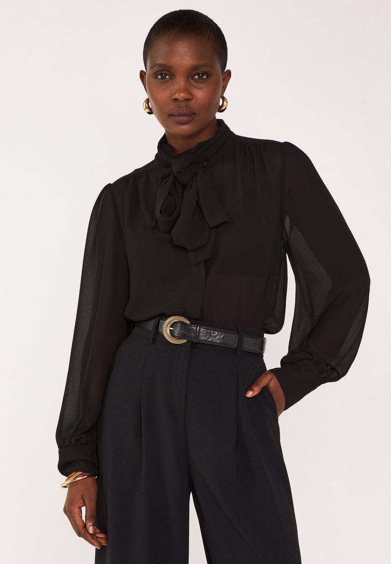 Blouse noire transparente avec un détail de col noué et des manches longues, associée à un pantalon noir taille haute et une ceinture noire texturée avec une boucle dorée.