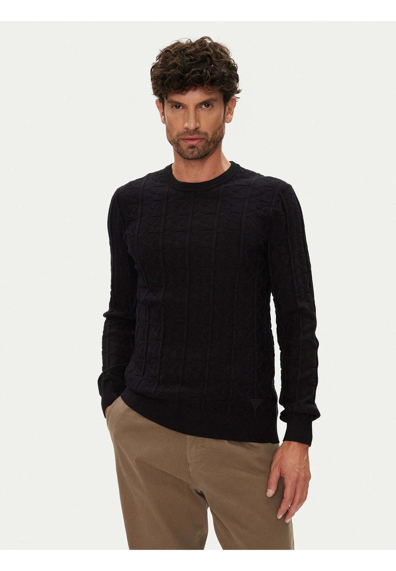 Marineblauer gestrickter Pullover mit strukturiertem Zopfmuster, rundem Ausschnitt und gerippten Bündchen; wird mit beigen Hosen getragen und besitzt ein Logo-Detail am Saum.