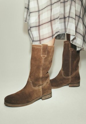 Braune Wildlederstiefel bis zur Mitte der Waden mit dekorativer Naht, getragen zu einem hellen karierten Rock.