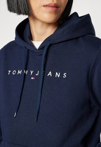Tmavě modrá mikina s kapucí z bavlny, na které je bíle vyšívaný text "TOMMYJEANS" a malá barevná logo akcentace blízko písmen.