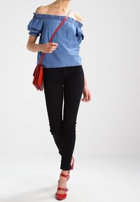 Top in denim blu off-the-shoulder con maniche a volant, abbinato a pantaloni neri aderenti e tacchi rossi con cinturini, mentre tiene una borsa rossa a tracolla.