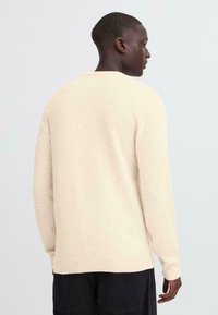 Beige, textured knit sweater with long sleeves and a round neckline. Features ribbed cuffs and hem. Worn from a side perspective.