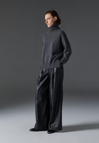 Tumši pelēks turtlenecks džemperis no mīkstas dzijas, apvienots ar platām melnām ādas biksēm. Modelis pozē, rokas ieliekot kabatās, pret neitrālu fona.