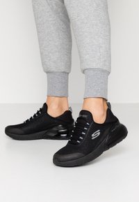 Czarne siatkowe sneakersy z teksturowaną cholewką, szarymi sznurowadłami i zaokrągloną podeszwą. Posiadają błyszczący czarny obcas i detal z logo po boku.