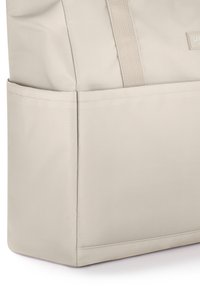 Beżowa torba na ramię z rogiem, przeszytymi szwami i szerokim, plecionym paskiem na ramię, ukazująca boczną kieszeń i część etykiety marki.