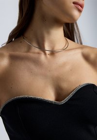 Fekete, pánt nélküli top ezüst díszítéssel a nyakvonal mentén. A modellen vékony fém choker nyaklánc van. Sima szövettextúra.
