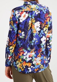 Långärmad blus i ett livligt blommönster på en mörkblå bakgrund. Har en krage och knäppte manschetter med en böjd nederkant.