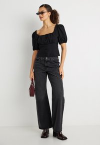 Femme aux cheveux bouclés portant un haut noir à manches ballon, un jean noir à jambes larges, des bottines à talons foncées, des lunettes de soleil noires, et tenant un petit sac à main bordeaux.