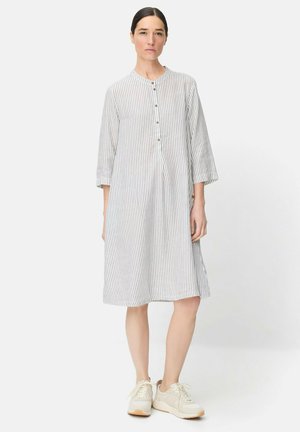 Kobieta stojąca w białej i szarej sukience w pionowe paski, sięgającej do kolan, z rękawami do łokcia i białych trampkach na jednolitym tle.