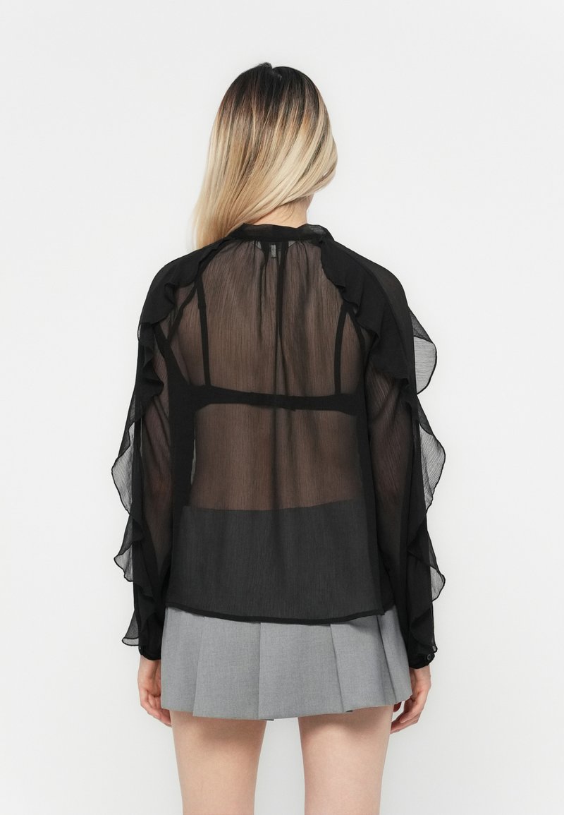 Blouse noire transparente avec des manches froncées, dotée d'un col montant et d'un dos transparent. Associée à une jupe plissée grise.