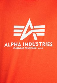 Oranges Baumwoll-T-Shirt mit einem großen weißen Logo, das ein stilisiertes "A" und horizontale Streifen zeigt. Der Text lautet "ALPHA INDUSTRIES" mit Standortdetails.